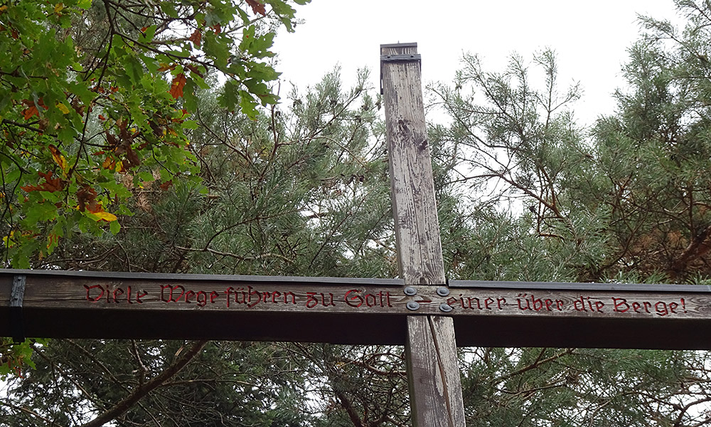 Kreuz mit Inschrift "Viele Wege führen zu Gott - einer über die Berge"