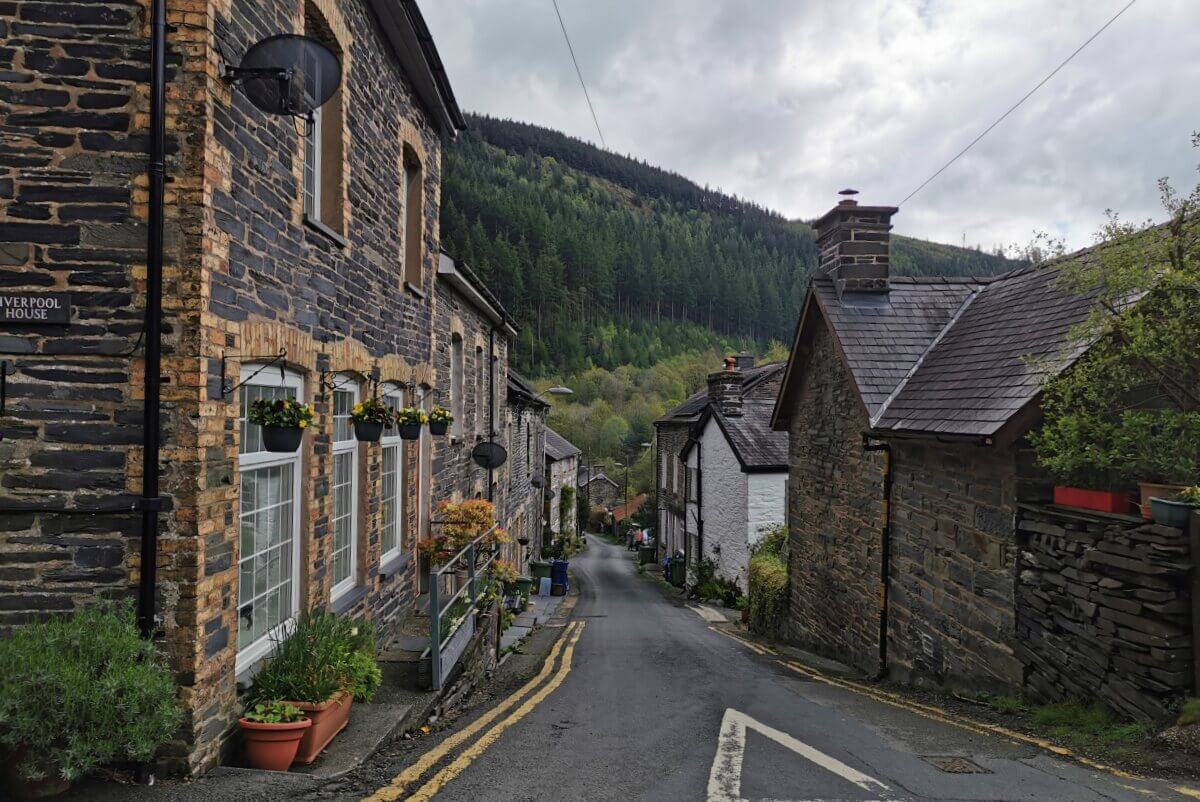 Eine schmale Straße gesäumt von Steinhäusern und Blumenkästen in einem kleinen Dorf, im Hintergrund erheben sich bewaldete Hügel unter einem bewölkten Himmel.