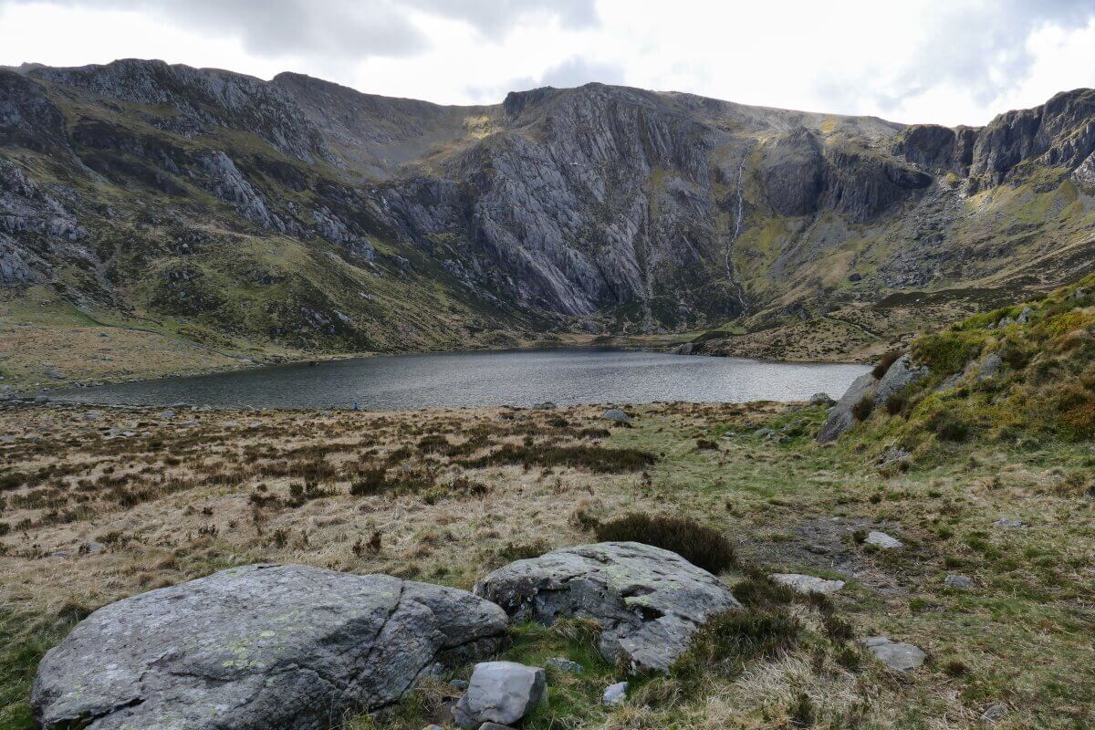 Ein kleiner Bergsee, umgeben von felsigen Hügeln und Grasland. Im Hintergrund erheben sich steile Felsen, m Vordergrund sind große Felsbrocken und Grasflächen zu sehen.