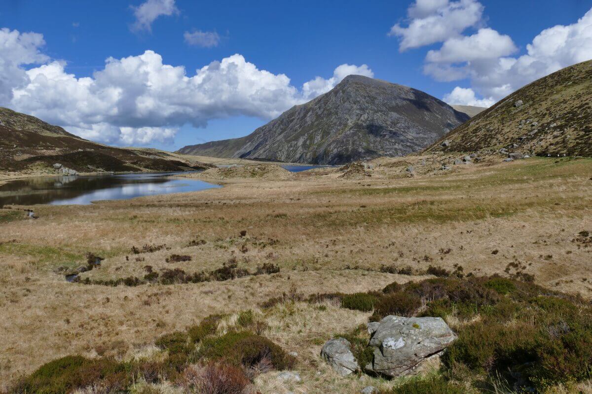 Ein grasbewachsenes Tal mit verstreuten Felsen führt zu einem ruhigen See, umgeben von sanften Hügeln und einem großen, schroffen Berg unter einem blauen Himmel mit weißen Wolken.