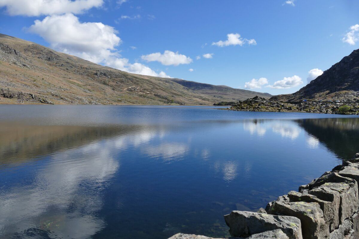 Ein ruhiger See, umgeben von felsigen Hügeln, mit blauem Himmel und weißen Wolken, die sich im Wasser spiegeln.