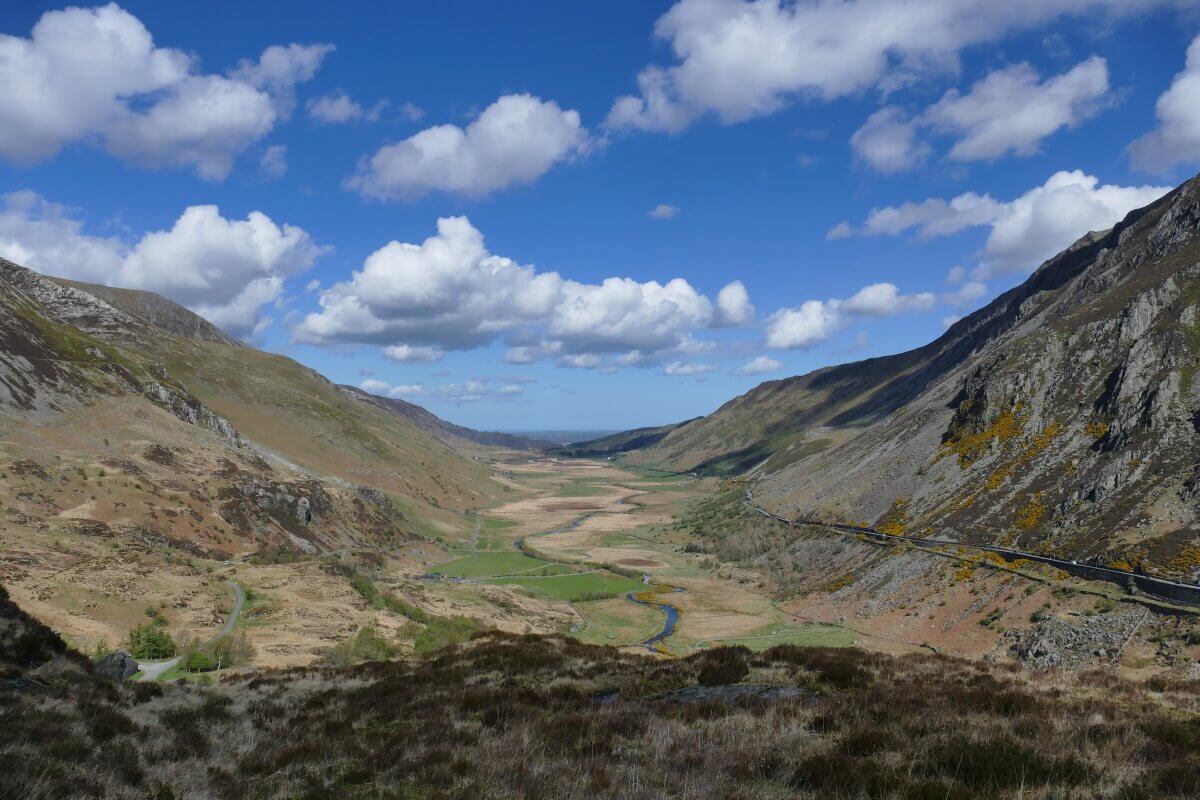 Ein weites Tal mit einem gewundenen Fluss, flankiert von steilen, felsigen Hügeln unter einem strahlend blauen Himmel mit vereinzelten weißen Wolken. Spärliches Gras und Flecken brauner Erde bedecken die Landschaft.