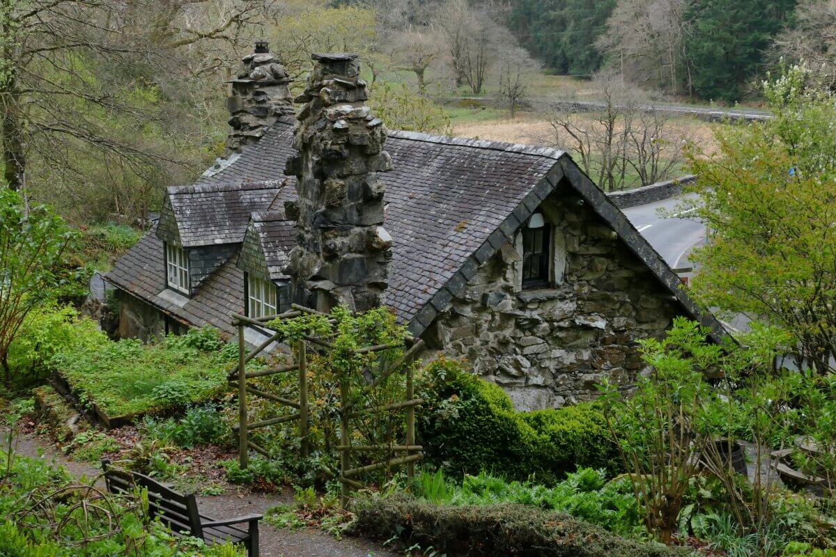 Ein rustikales Steinhaus mit zwei hohen Schornsteinen liegt eingebettet zwischen üppigem Grün und Bäumen, mit einem Gartenweg und einer Holzbank im Vordergrund.