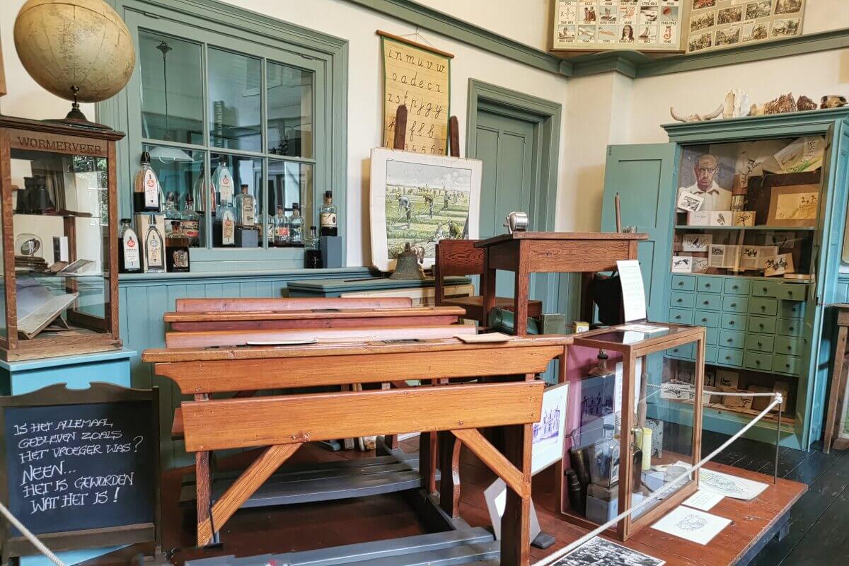 Ein altes Klassenzimmer mit Holztischen, hellblauen Türen und Schrank, einem großen Globus, alten Unterrichtsmaterialien, Postern und Vitrinen voller historischer Schulgegenstände. 