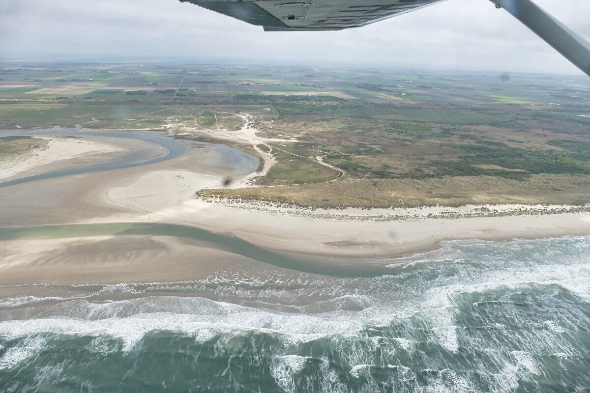 Luftaufnahme einer sandigen Küste mit brechenden Wellen, einem Fluss, der sich durch die Landschaft schlängelt, und Ackerland im Hintergrund. Oben im Bild ist ein Teil eines Flugzeugflügels zu sehen. 