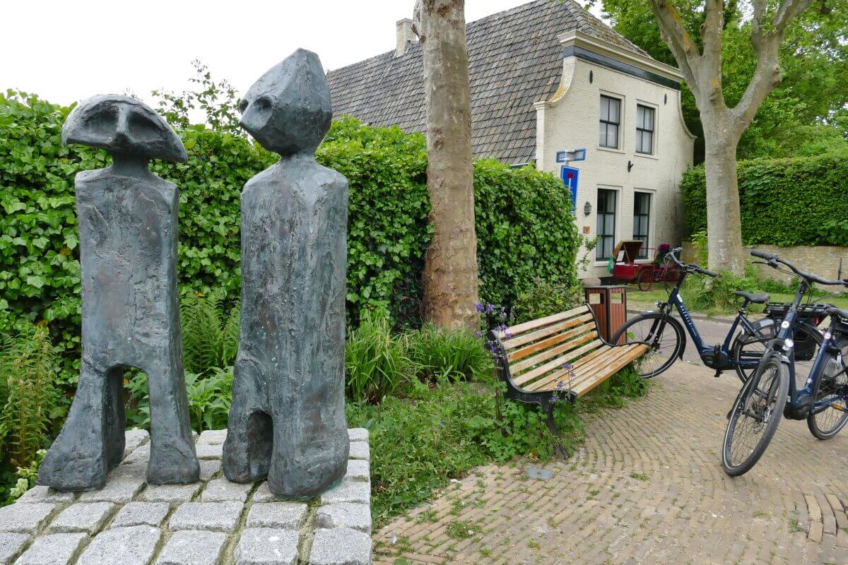 Zwei Steinfiguren, die Sommeltjes, stehen auf einem Steinsockel neben einer Bank und zwei Fahrrädern, im Hintergrund eine Hecke, Bäume und ein helles Haus auf einem Kopfsteinpflasterweg. 