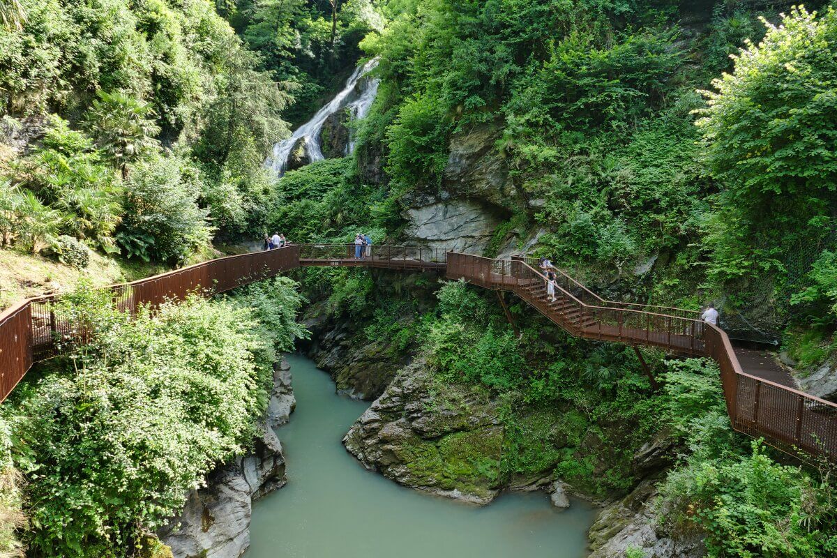 Ein Steg schlängelt sich durch üppig grüne Felsen über einem schmalen Fluss, mit einem Wasserfall im Hintergrund. 