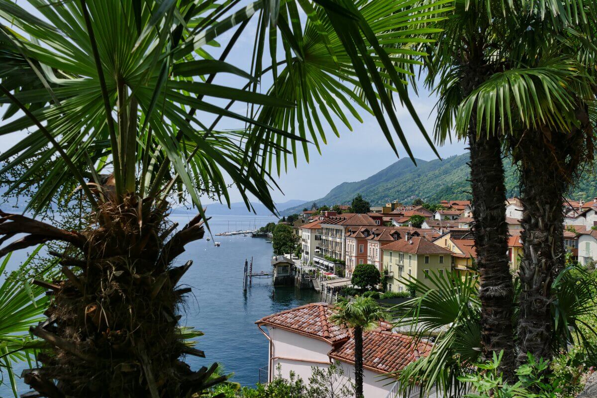 Blick auf ein Dorf mit bunten Gebäuden am Wasser, eingerahmt von hohen Palmen im Vordergrund, mit Bergen und einem ruhigen See.
