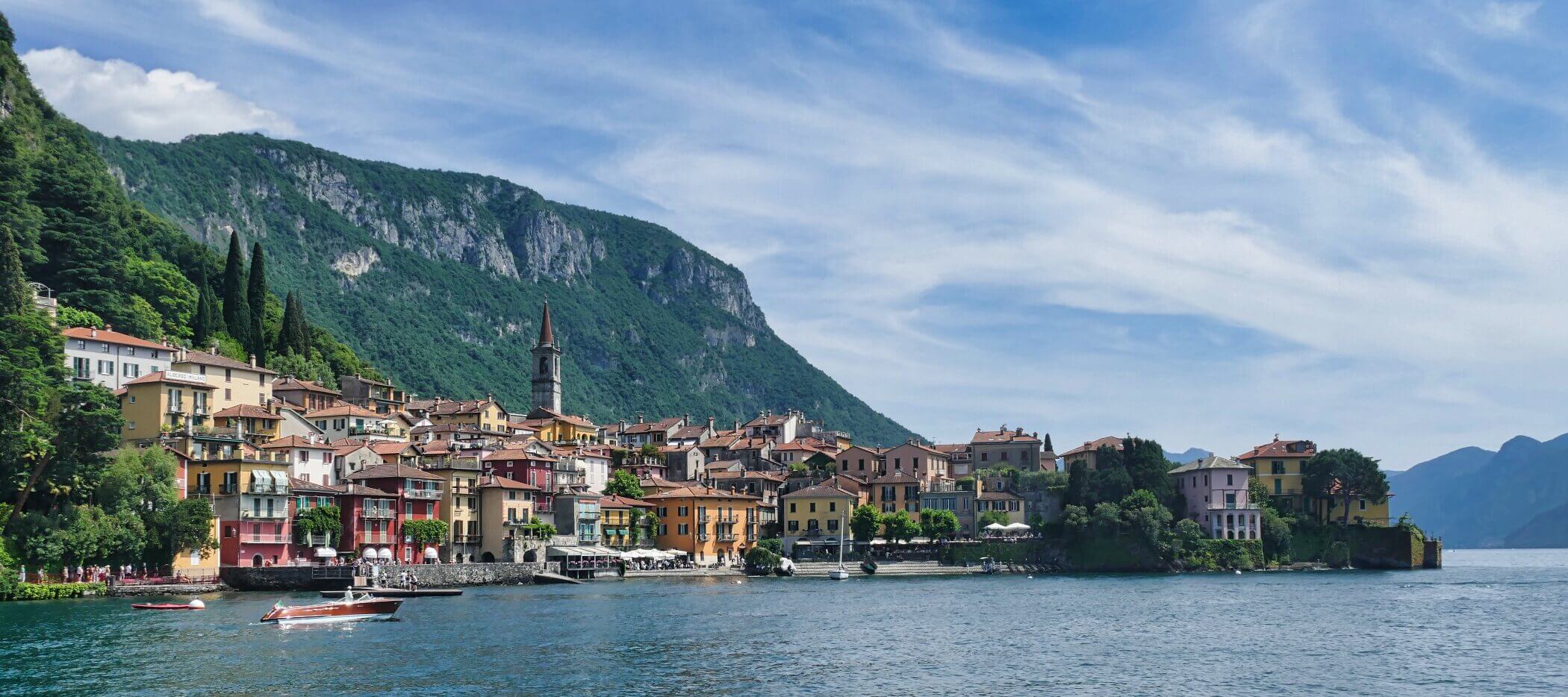 Bunte Gebäude säumen das Ufer eines Dorfes am See, umgeben von grünen Hügeln und Bergen unter einem teilweise bewölkten Himmel, mit Booten auf dem blauen Wasser im Vordergrund.