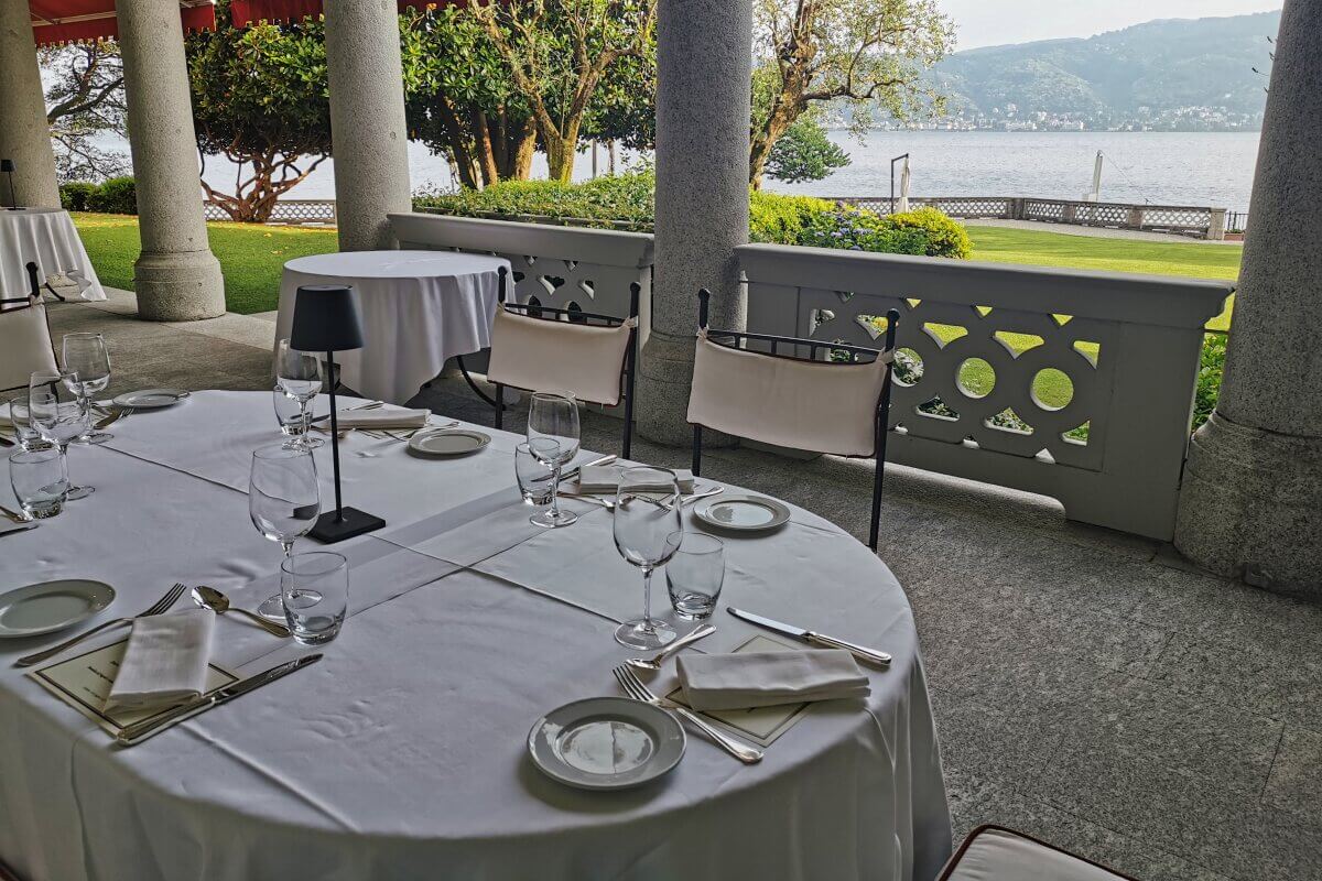 Ein mit Gläsern gedeckter Tisch im Freien, mit Blick auf einen See und die Berge, beschattet von Säulen und Bäumen. 