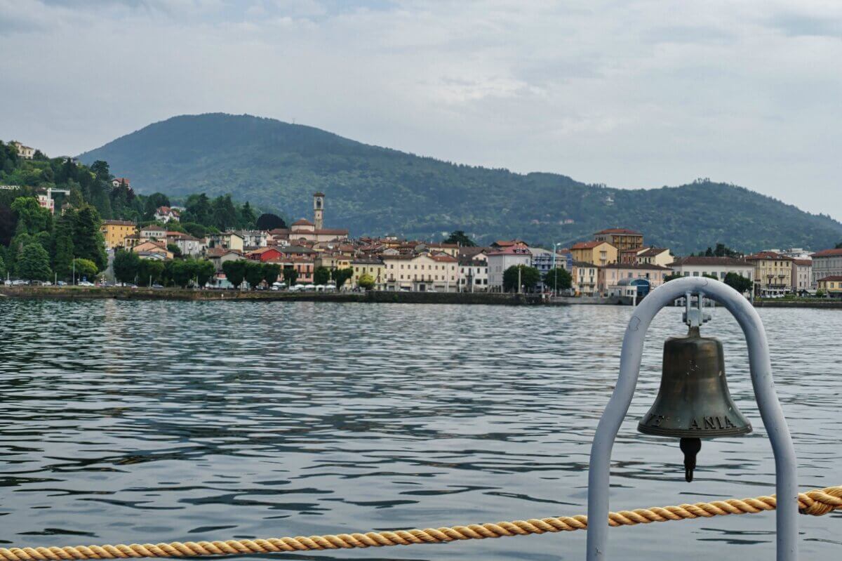 Eine Stadt am Seeufer mit Bergen im Hintergrund, gesehen von einem Schiff mit einer Glocke im Vordergrund. 