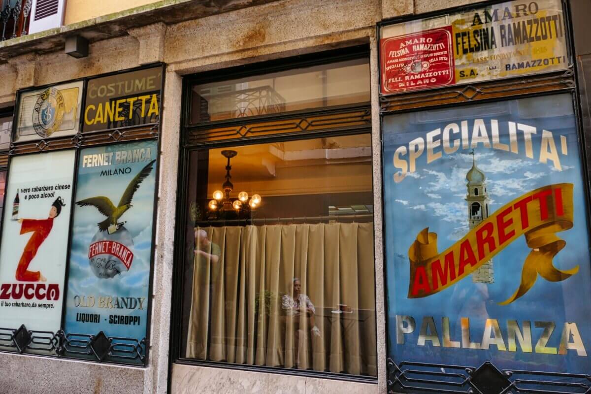 Schaufenster mit alten italienischen Schildern und Plakaten, in denen sich ein zwei Personen spiegeln. 