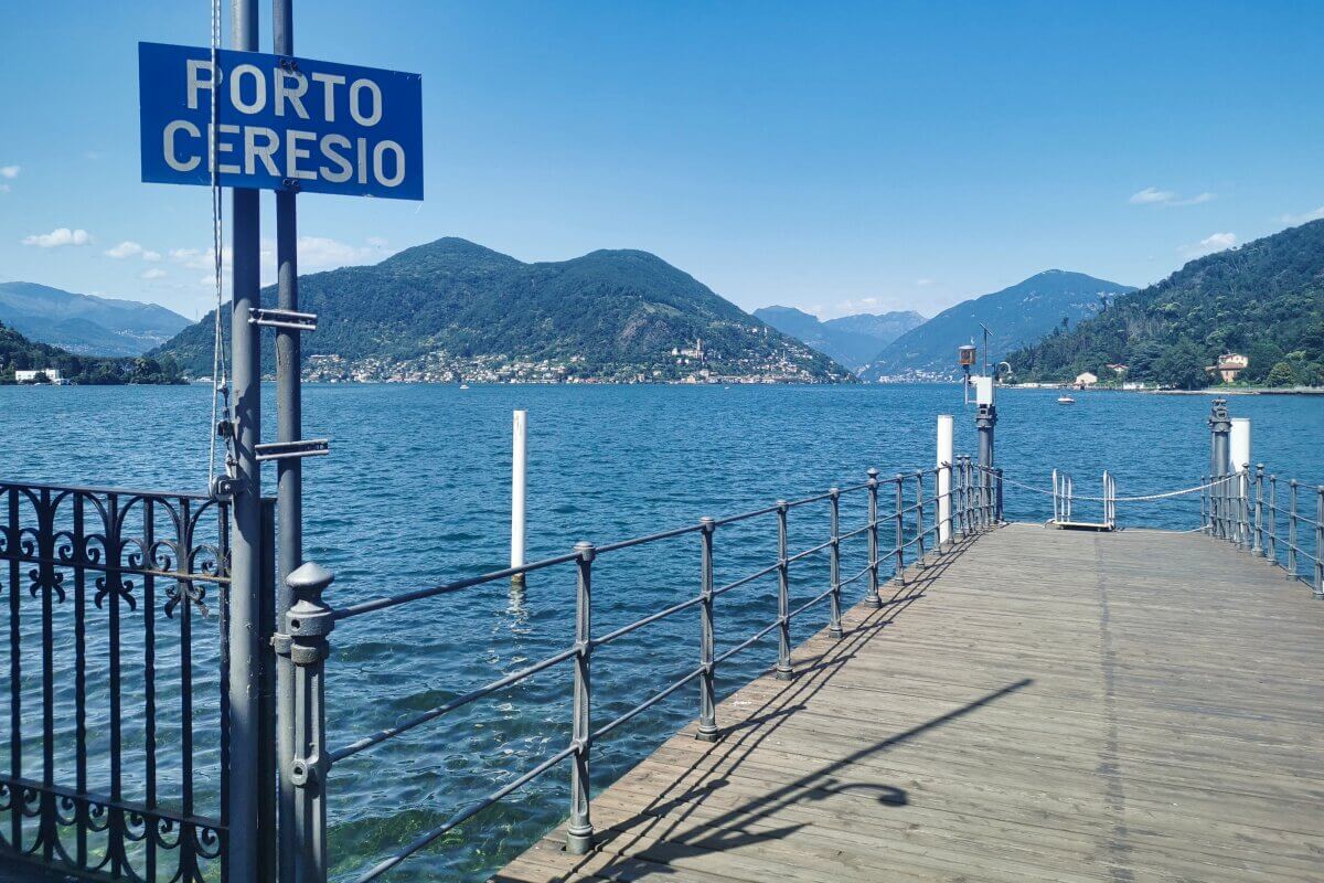 Holzsteg mit Porto Ceresio-Schild, mit Blick auf den Luganer See und Berge unter einem klaren blauen Himmel. 