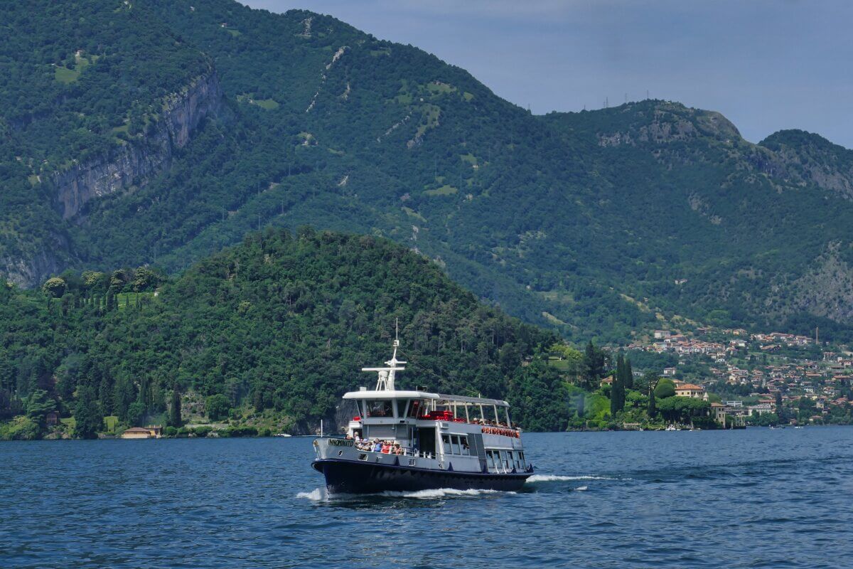 Ein Boot fährt auf dem Luganer See mit grünen Bergen und einem Dorf im Hintergrund unter einem blauen Himmel. 