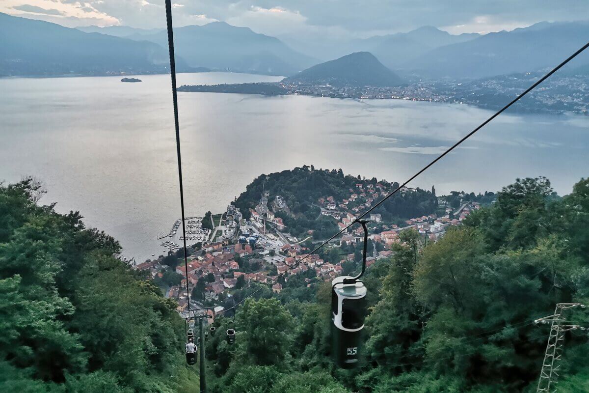 Eine Seilbahn führt über einen üppig bewachsenen Hügel, von dem aus man in der Abenddämmerung einen Blick auf eine Stadt am Lage Maggiore und die umliegenden Berge hat. 