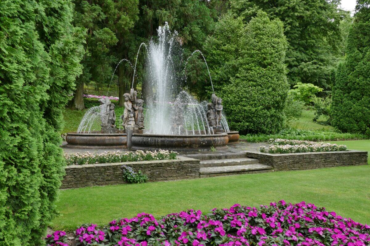 Ein Steinbrunnen mit Statuen und sprühendem Wasser, umgeben von üppigem Grün und violetten Blumen. 