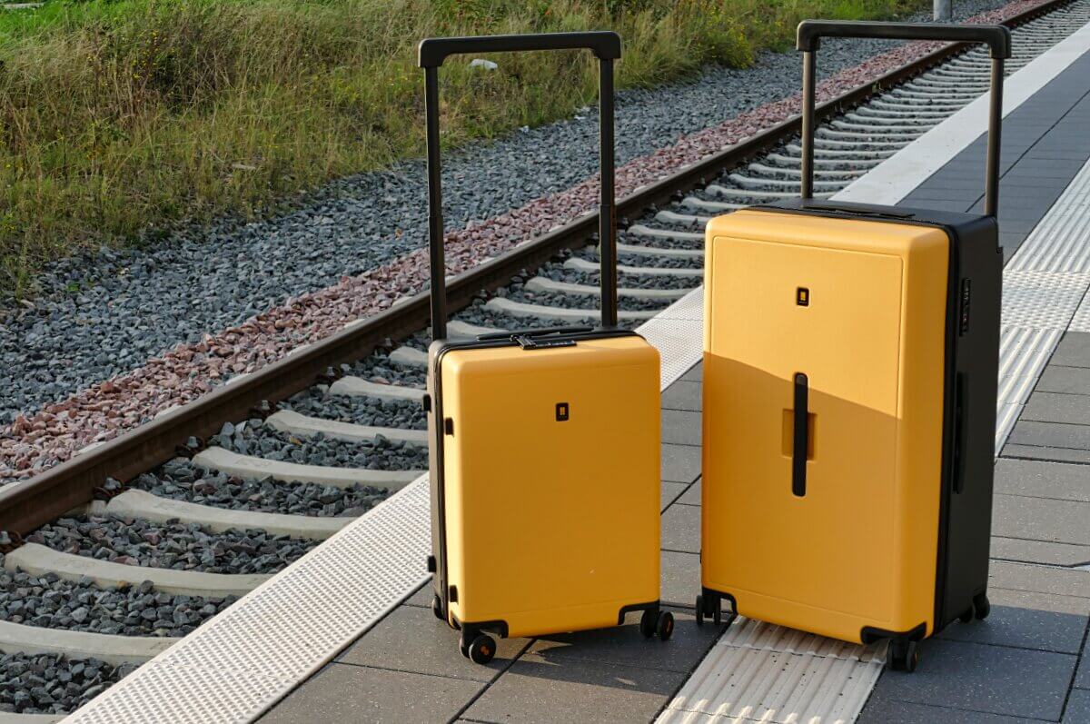 Zwei gelbe Koffer mit schwarzen Griffen stehen auf einem Bahnsteig neben den Gleisen.