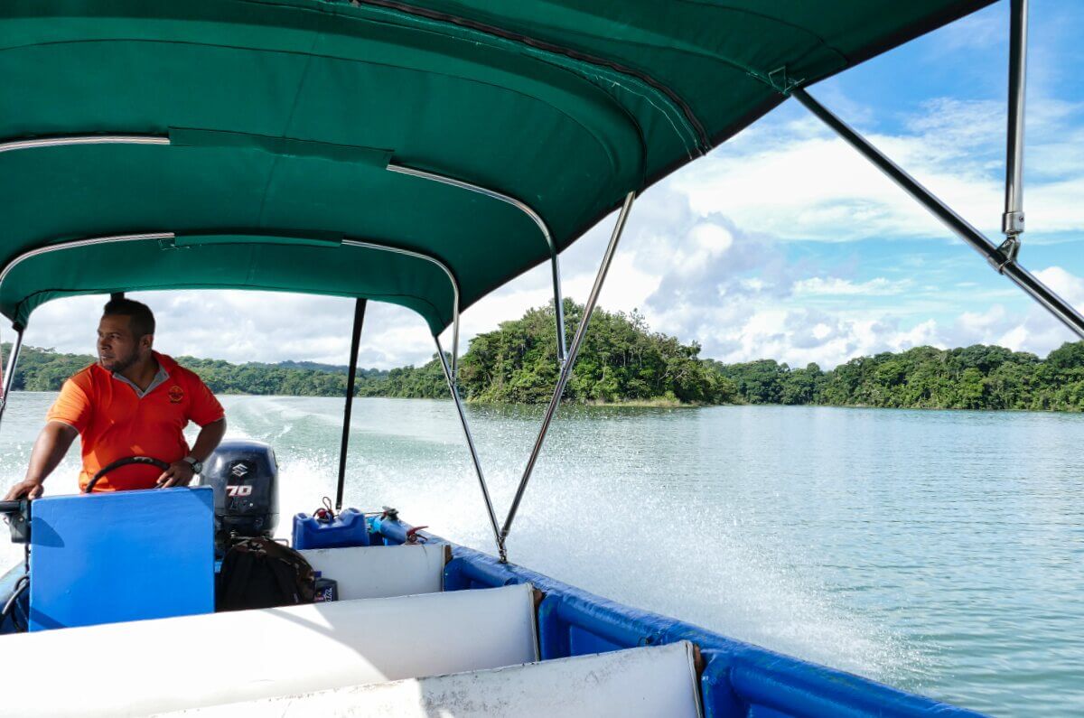 Ein Mann steuert ein kleines Motorboot auf einem ruhigen Fluss mit grünen Bäumen und blauem Himmel im Hintergrund.