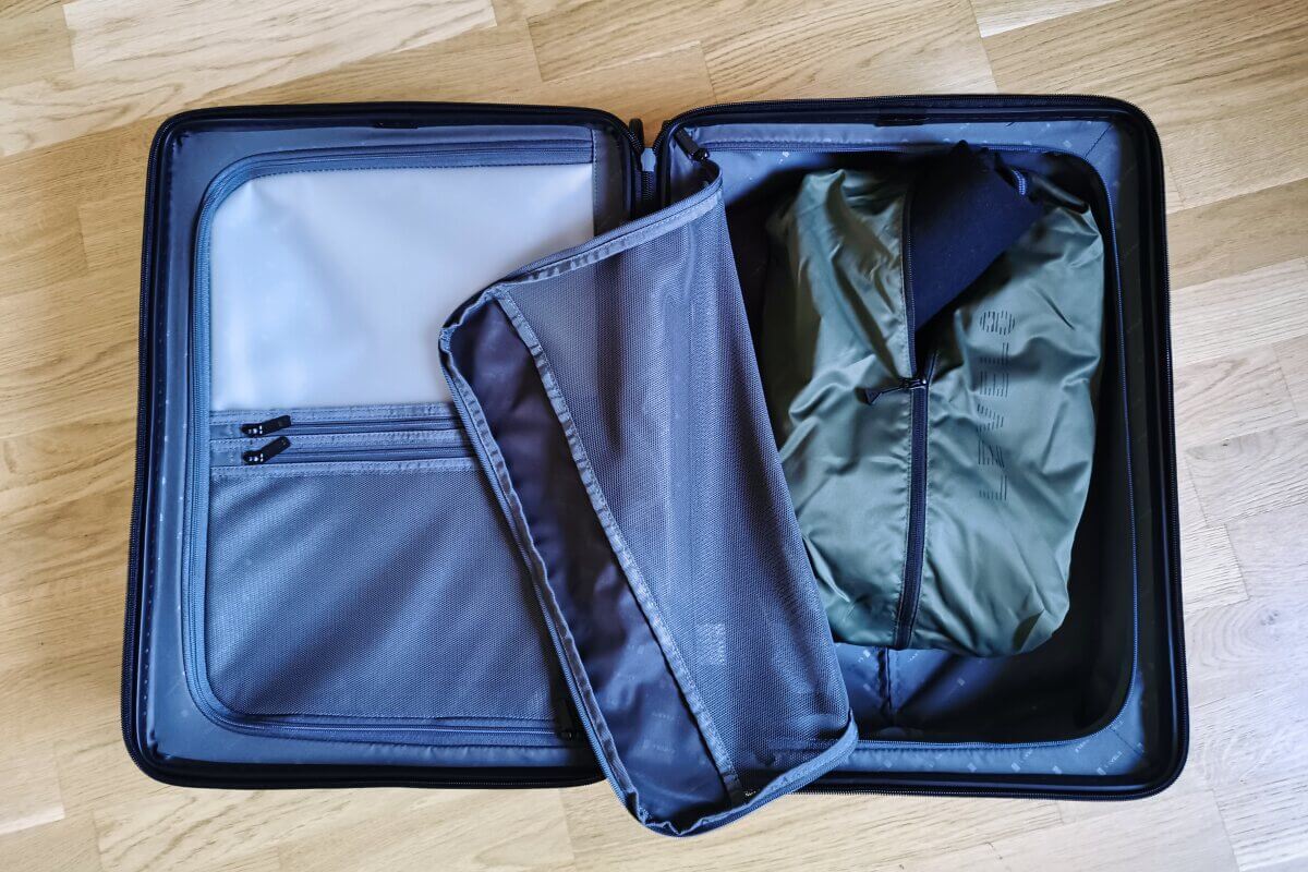 Ein offener Koffer mit Netztrennwänden und einer gepackten grünen Tasche darin, auf einem Holzboden.