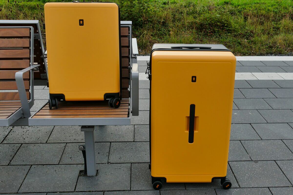 Zwei gelbe Koffer, einer auf einer Bank und einer auf dem Boden, in einem gepflasterten Außenbereich.