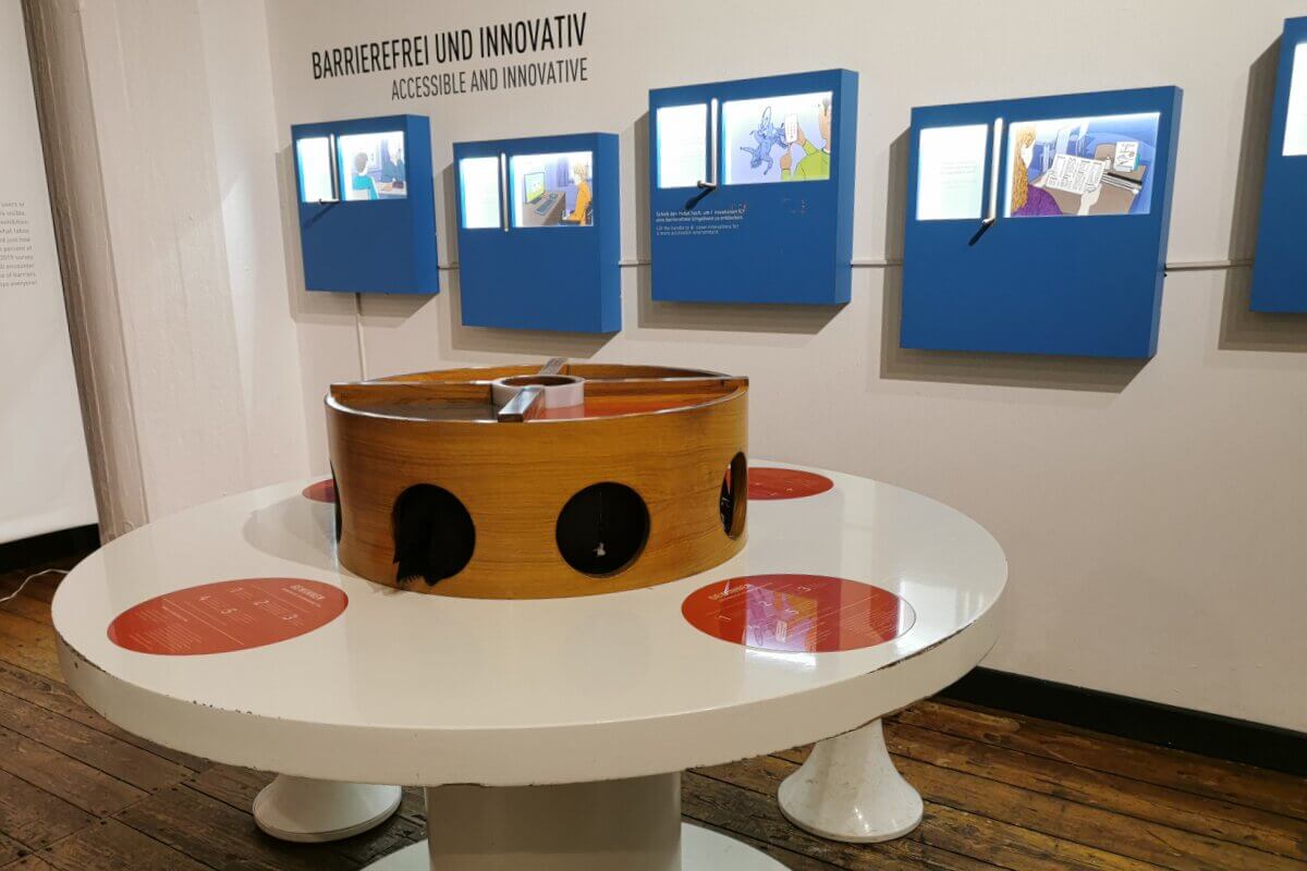 Exponat mit blauen interaktiven Tafeln an der Wand und einem runden taktilen Ausstellungstisch mit Handlöchern.
