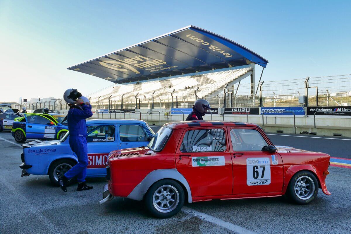 Zwei Fahrer in Rennanzügen stehen neben Oldtimern auf einer Rennstrecke, mit leeren Tribünen im Hintergrund.