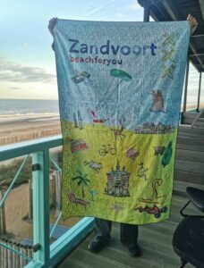 Person, die ein buntes Zandvoort-Strandtuch mit Illustrationen hochhält, auf einem Balkon mit Blick auf das Meer.