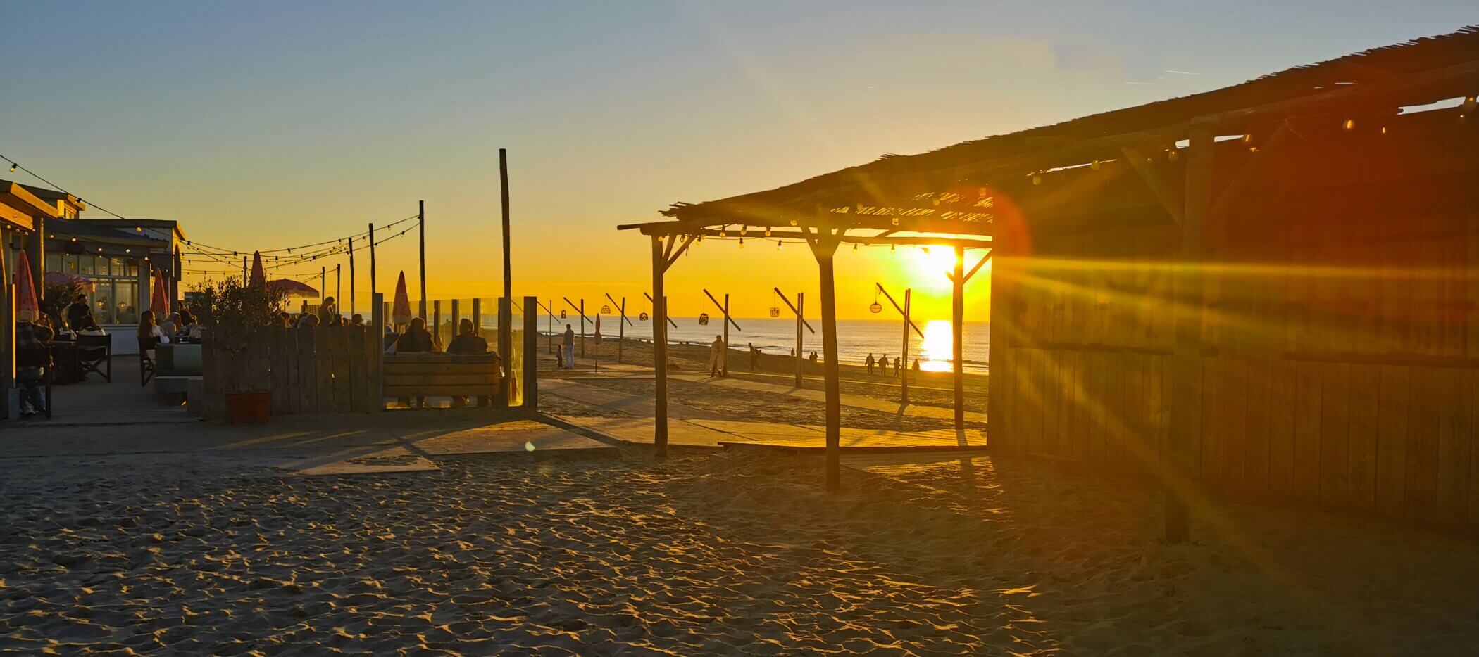 sonnenuntergang-zandvoort Strandcafé bei Sonnenuntergang mit Sonnenstrahlen, Holzhütten, sitzenden Menschen und Fahnen am Meer.