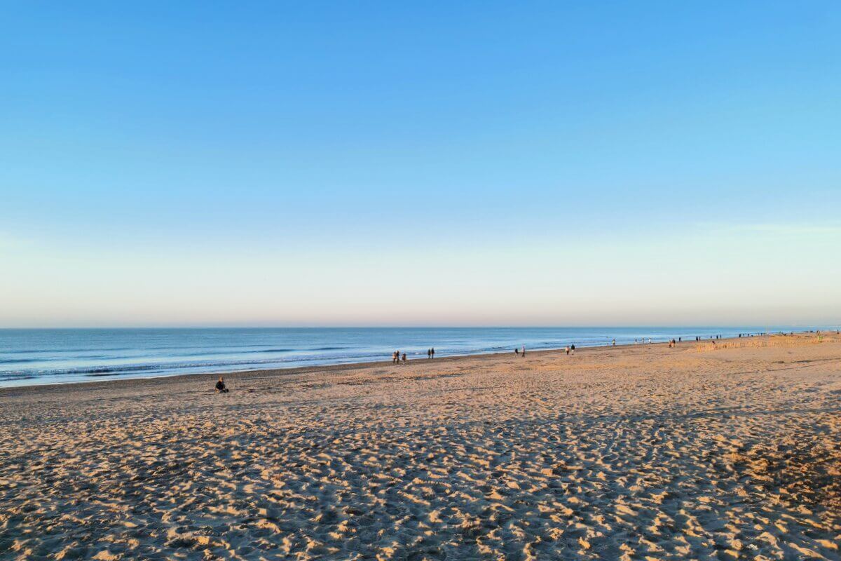 Sandstrand mit ruhigen Wellen, wenige Menschen, die bei Sonnenuntergang unter blauem Himmel am Strand spazieren gehen.