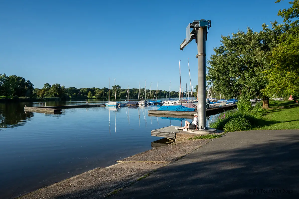 Kleiner Jachthafen mit angedockten Segelbooten, ruhiges Wasser, Grünflächen und eine Person, die an einem sonnigen Tag am Ufer sitzt.