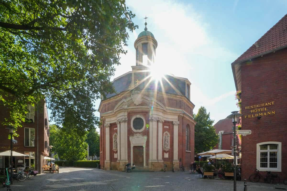 Das Sonnenlicht scheint hinter einer historischen Barockkirche auf einem kopfsteingepflasterten Platz, der von Bäumen und Gebäuden umgeben ist.
