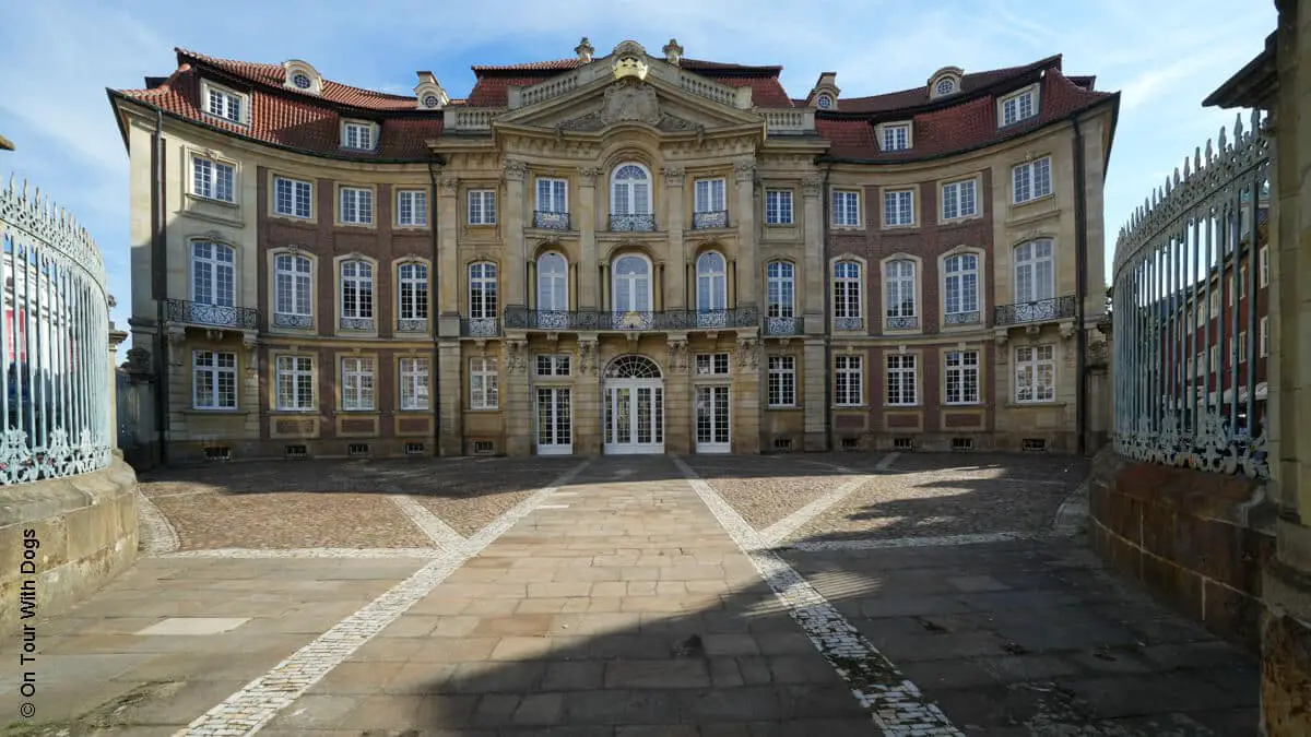 Ein prächtiges, historisches Gebäude mit Bogenfenstern, rotem Dach und verzierter Fassade, das von einem umzäunten Innenhof aus zu sehen ist.