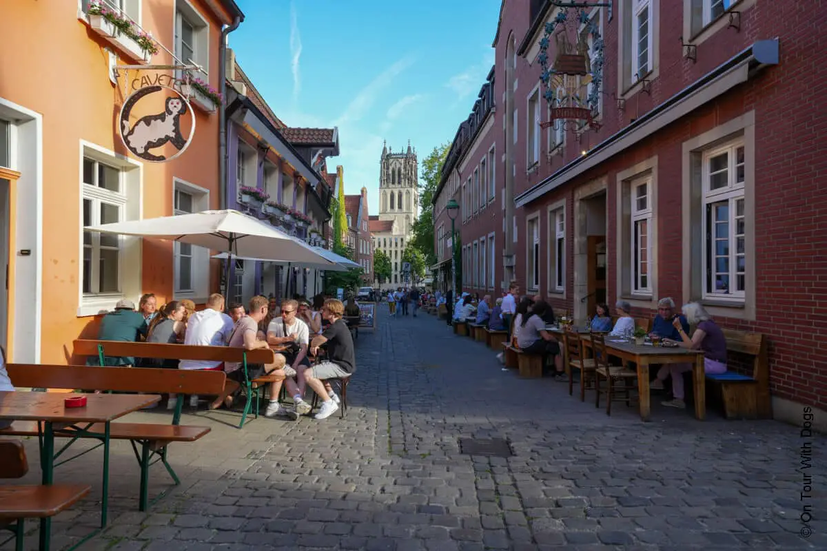Menschen speisen im Freien entlang einer Kopfsteinpflasterstraße mit einer Kirche am Ende der Straße.