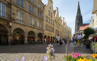 Menschen gehen auf einer Kopfsteinpflasterstraße, die von Geschäften und Blumen gesäumt ist, mit einer Kirchturmspitze im Hintergrund.