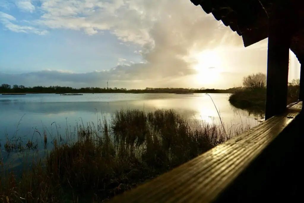 Ein Blick auf einen ruigen See bei Sonnenuntergang, gesehen von einem hölzernen Unterstand mit Schilf im Vordergrund.