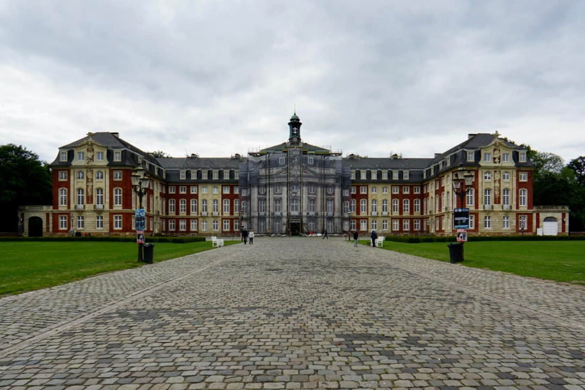 Historisches Schloss mit einem Kopfsteinpflasterweg, symmetrischen Flügeln und einem wolkenverhangenen Himmel.