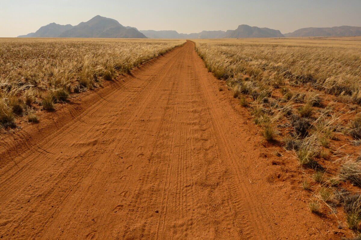 Eine rote, unbefestigte Straße in Namibia führt durch trockenes Grasland zu fernen Bergen unter einem klaren Himmel.
