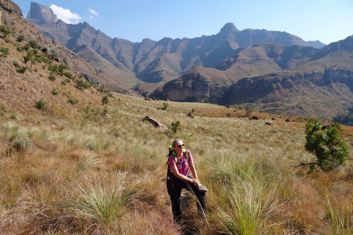 Eine wandernde Frau steht im hohen Gras mit Bergen im Hintergrund unter einem klaren Himmel.