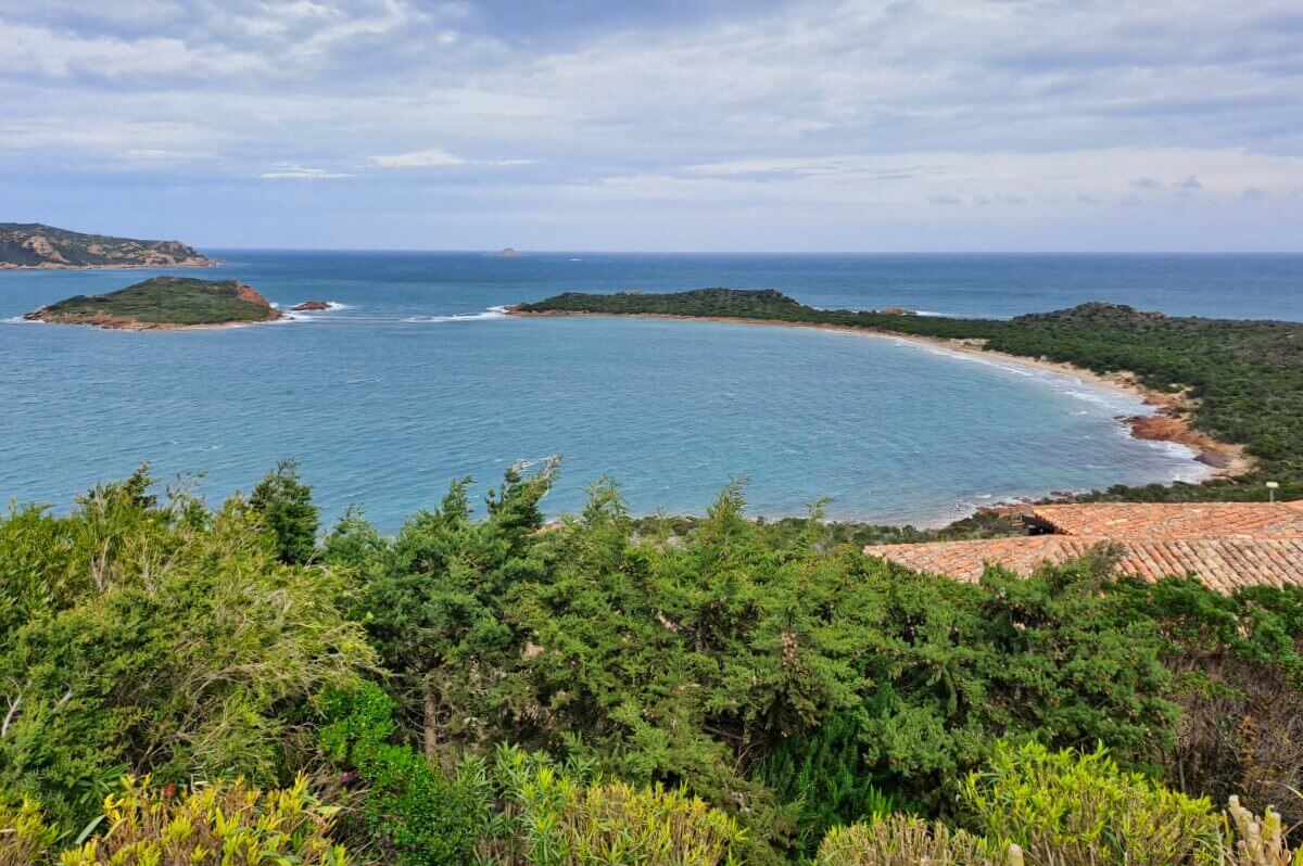 Eine geschwungene Bucht mit Sandstrand, grünen Bäumen und blauem Wasser unter einem bewölkten Himmel.