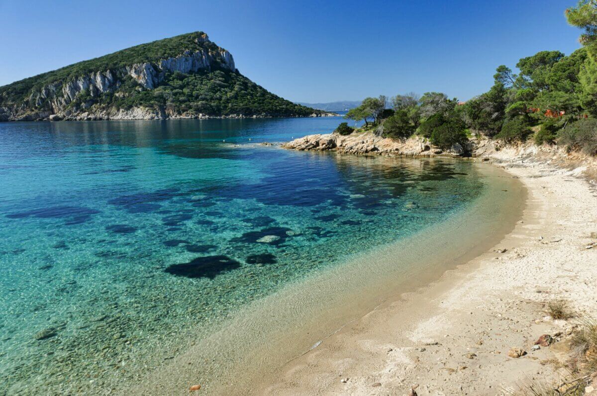 Klares türkisfarbenes Wasser und ein Sandstrand mit einer felsigen, grünen Insel und blauem Himmel im Hintergrund.