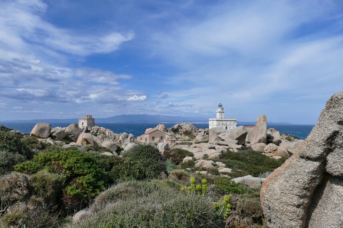 Felsige Landschaft mit grünen Sträuchern, alten Steingebäuden und einem weißen Leuchtturm unter einem teilweise bewölkten Himmel.