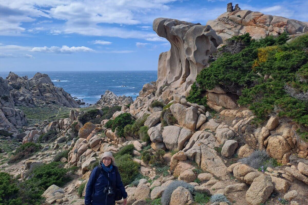 Eine Person wandert zwischen felsigen Klippen und grüner Vegetation am Meer unter einem teilweise bewölkten Himmel.