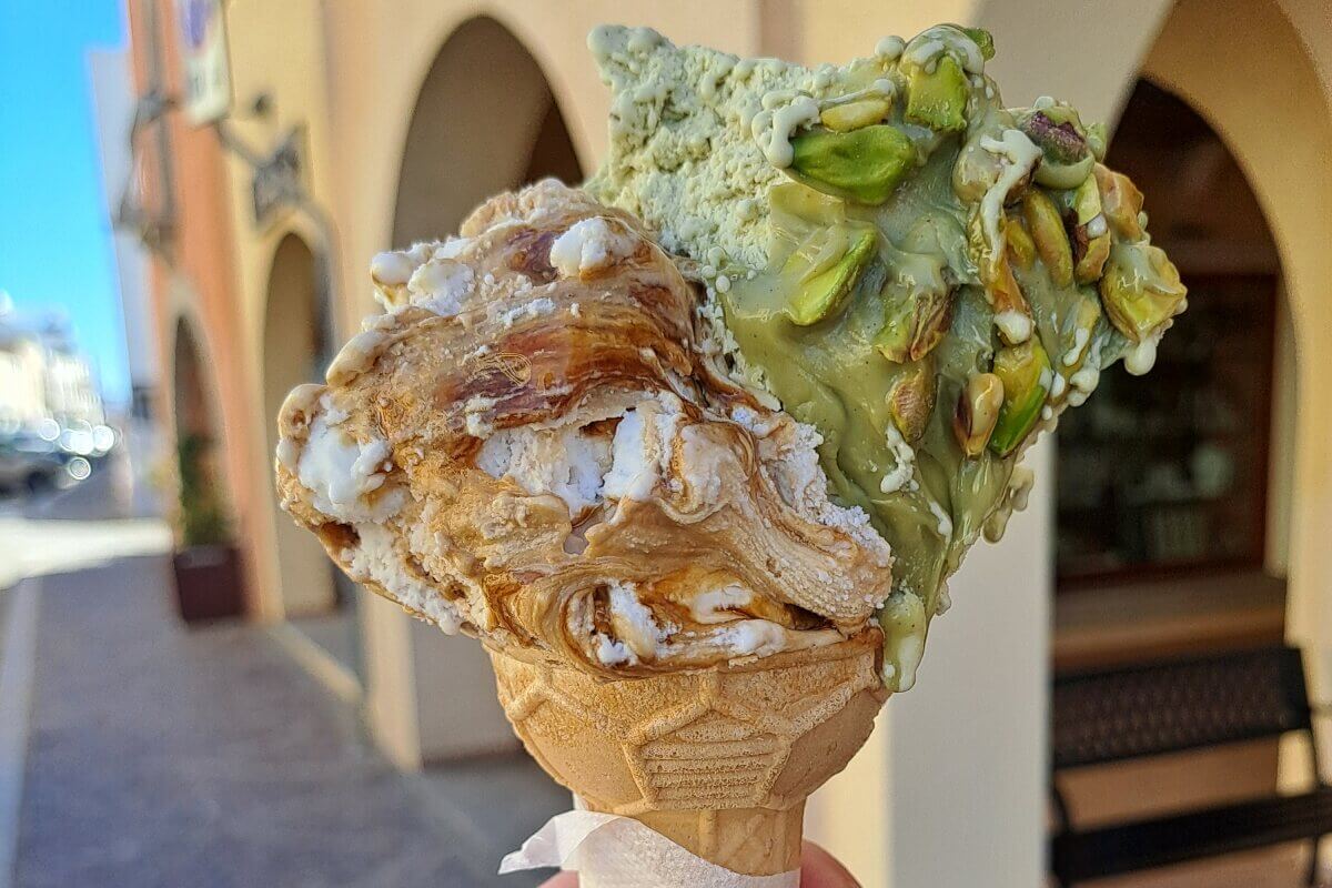 Eine Hand hält eine Waffel mit zwei Kugeln Eis, eine mit Pistazie und eine mit Karamell, in einer sonnigen Straßenumgebung.