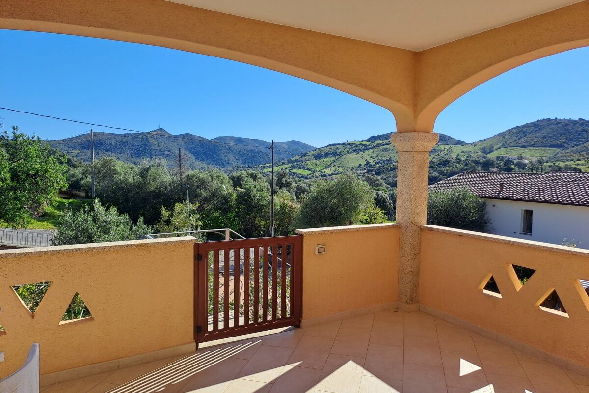 Sonnige Terrasse mit gefliestem Boden und Holztor mit Blick auf grüne Hügel und ein Haus mit Ziegeldach.