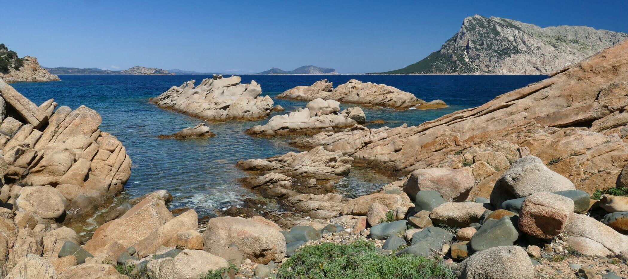 sardinien-punta-molara-2 Felsenküste mit klarem, blauem Wasser, verstreuten Felsen und einem entfernten Berg unter strahlend blauem Himmel.
