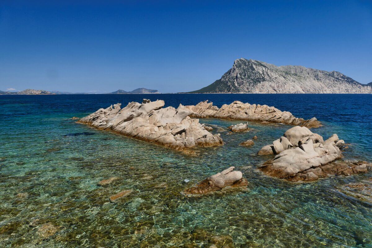 Felseninseln im klaren blauen Wasser mit einem großen Berg und blauem Himmel im Hintergrund.