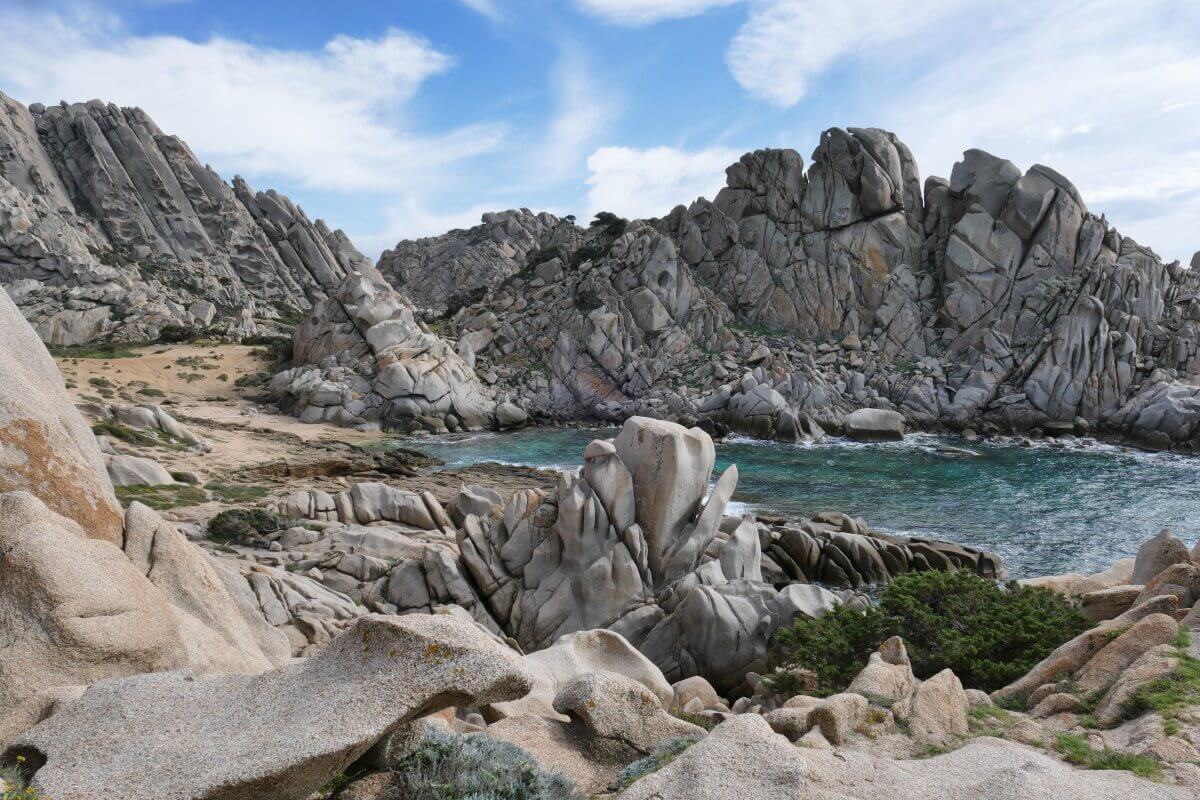 Felsige Küstenklippen und Felsbrocken säumen eine kleine Bucht mit blauem Wasser unter einem teilweise bewölkten Himmel.