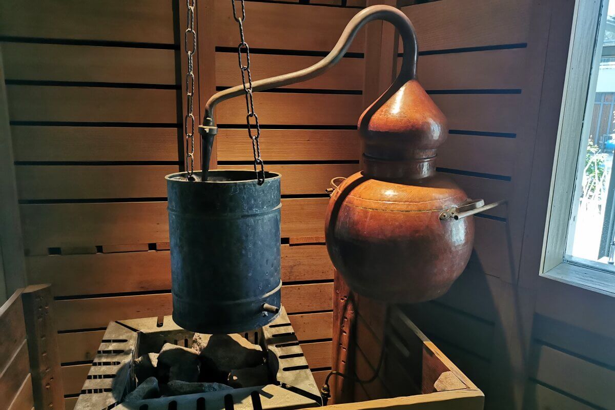 In einem holzgetäfelten Raum hängt ein Metalltopf über Steinen neben einer Destillationsvorrichtung aus Kupfer.
