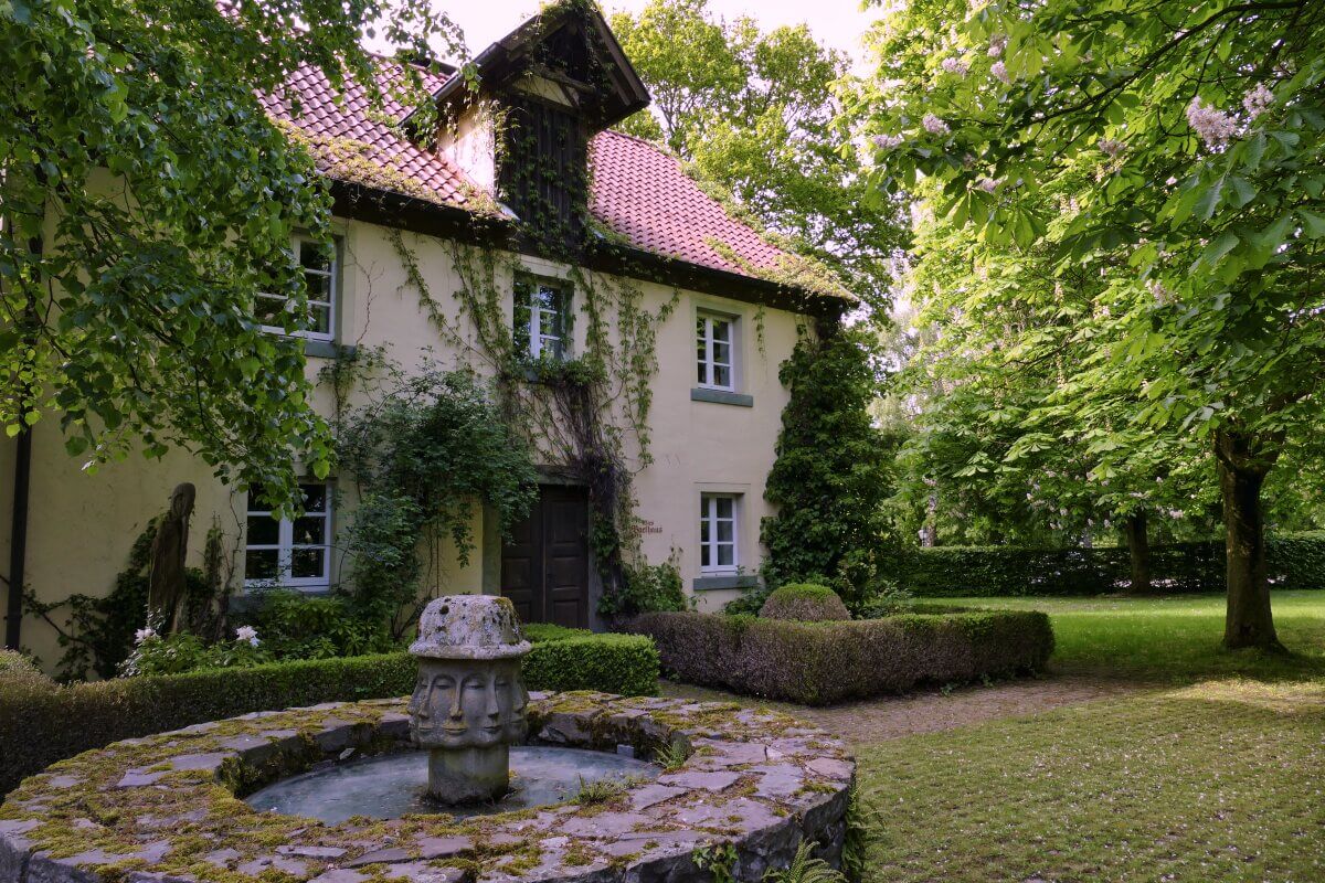 Vor einem gelben, mit Weinreben bewachsenen Haus, das von üppig grünen Bäumen und Sträuchern umgeben ist, steht ein Steinbrunnen.