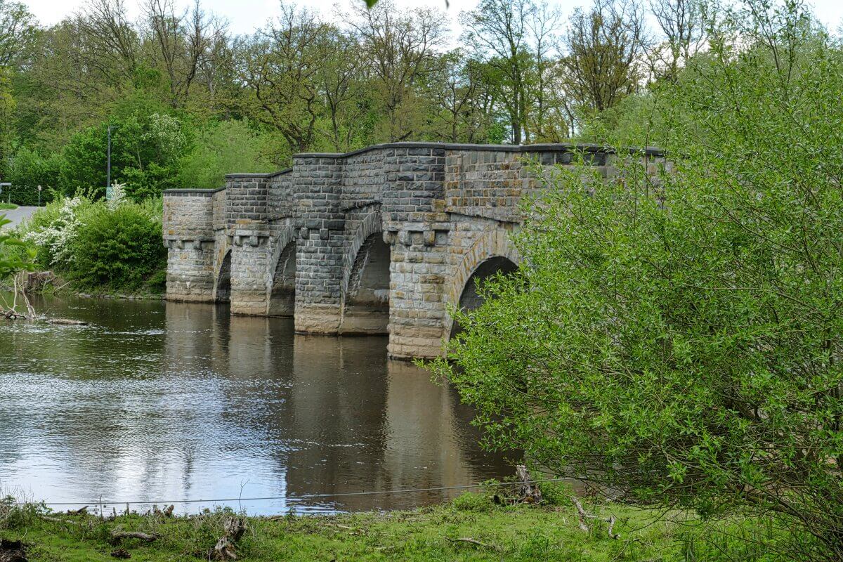 Eine Steinbogenbrücke überspannt einen ruhigen Fluss, umgeben von grünen Bäumen und Sträuchern.